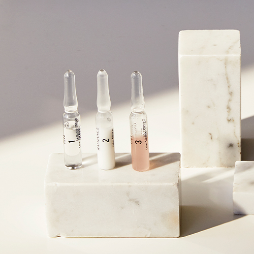 De drie verschillende ampullen van de Diamond Instant Glow behandeling liggend op een spiegelend oppervlak met een paarse lichtinval.