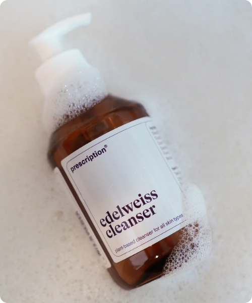 Een pompflacon van Prescription edelweiss cleanser ligt op een lichtgekleurd oppervlak en is bedekt met wit schuim