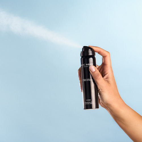 Een close up van een hand die de Marc Inbane SPF 50 Setting Mist invisible vernevelt waarbij de lichte neveltextuur goed zichtbaar is tegen een blauwe achtergrond
