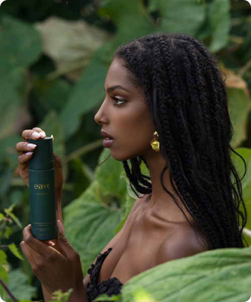 Een vrouw met lang gevlochten haar wordt van opzij getoond terwijl ze een donkergroene flacon van EAVE 'I AM RADIANT' vasthoudt. Ze staat in een natuurlijke omgeving met grote, groene bladeren op de achtergrond.