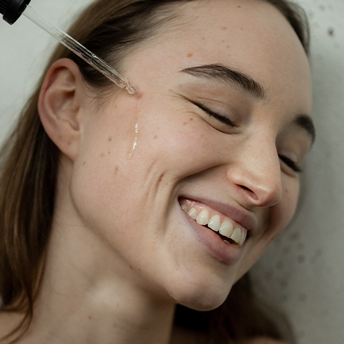 Een close-up portret van een lachende jonge vrouw met sproetjes, die haar ogen half dichtknijpt. Een glazen pipet van Likami komt van bovenaf in beeld en laat een druppel van de amberkleurige Skin Ferment Essence op haar wang vallen.