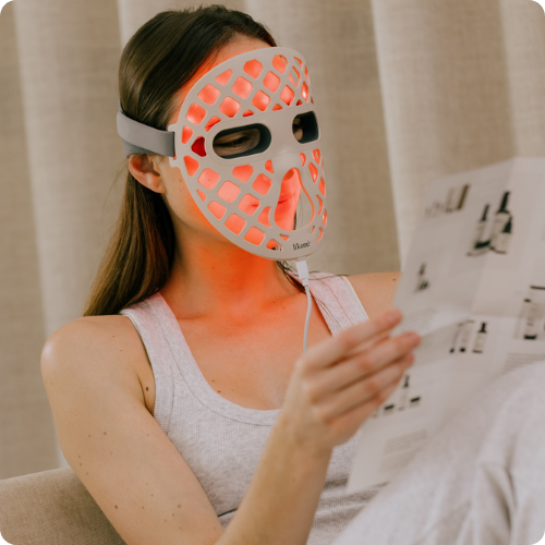 Een vrouw op de bank met het witte Likami LED-masker dat een rode gloed verspreidt. Ze leest gefocust in een brochure. De afbeelding heeft afgeronde hoeken.