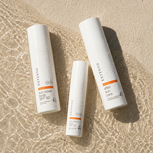 Een productfoto van drie witte Extenso zonneverzorgingsproducten – twee flessen zonnebrandcrème voor het lichaam (SPF 50) en één fles after sun lotion – liggend op een natte, zandkleurige ondergrond met rimpelingen in het water.
