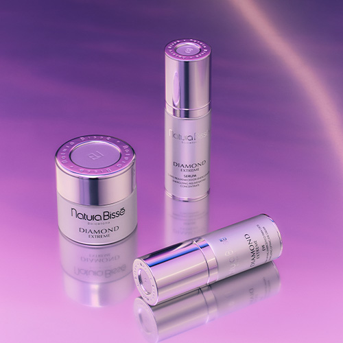 Een compositie van drie Natura Bissé Diamond Extreme producten — een pot, een staande flacon en een liggende flacon — op een reflecterend paars oppervlak.