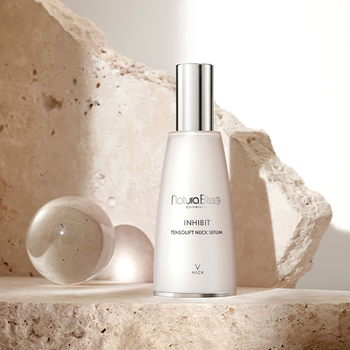 Een sfeerbeeld van het Natura Bissé Inhibit Tensolift Neck Serum op een stenen plateau, geflankeerd door een glazen bol en rustieke rotswanden in warm licht.