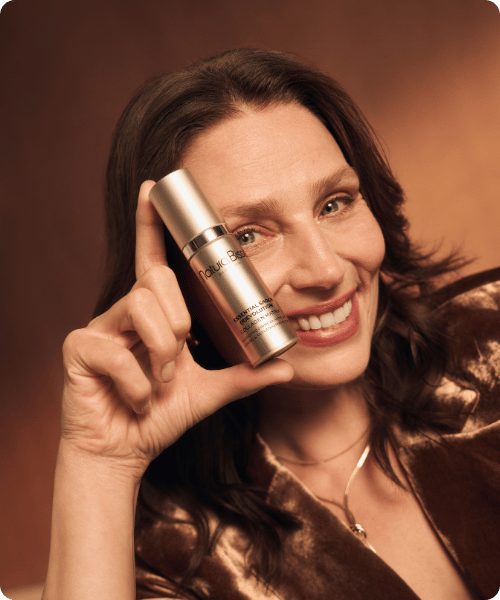 Een lachende vrouw met golvend bruin haar houdt een zilveren sprayflacon van Natura Bissé Essential Shock Intense Complex vast bij haar slaap. Ze draagt een bruine, glanzende fluwelen blazer tegen een warme, bruine achtergrond.