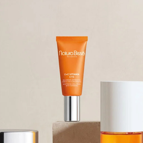 De oranje tube oogcrème rechtopstaand op een stenen blok in een minimalistische, warm verlichte omgeving.