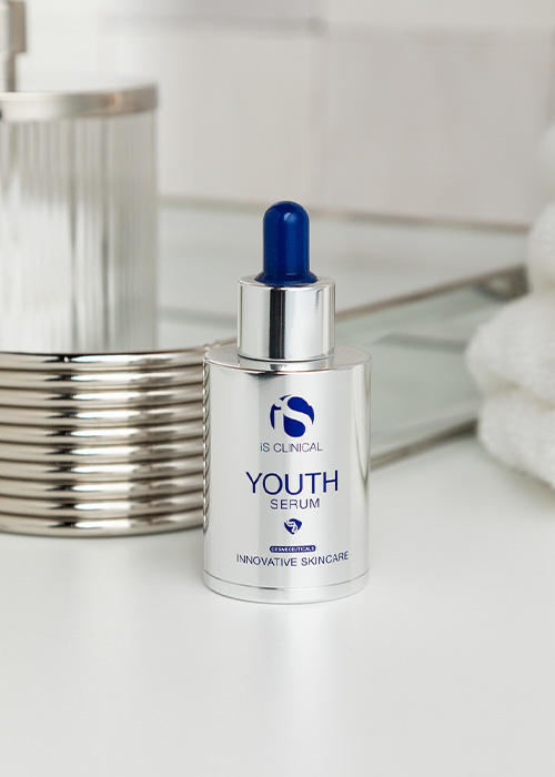 Een flesje iS Clinical Youth Serum staand op een witte ondergrond met een zilveren spiegel en handdoeken op de achtergrond.
