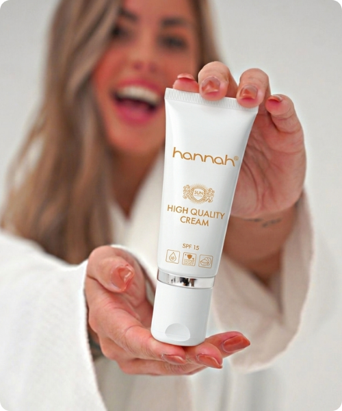 Een vrouw in een witte badjas houdt een tube hannah High Quality Cream SPF15 dicht bij de camera, terwijl zij lachend op de achtergrond vervaagd is.