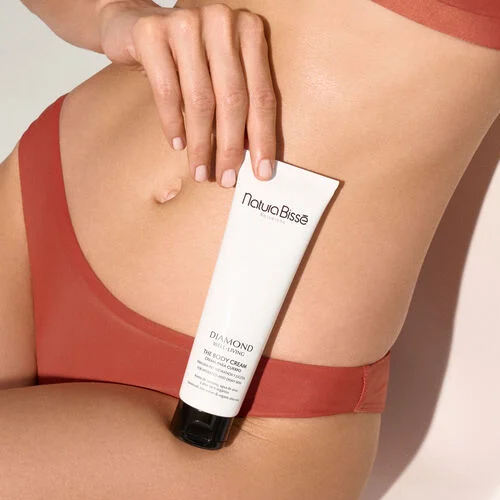 Een close-up foto van een hand die de Natura Bissé The Body Cream tube vasthoudt tegen een blote buik en dij, gekleed in een terra-cotta kleurige bikinislip. De focus ligt op het product en de huid, met een zachte, warme verlichting.