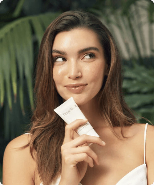 Een vrouw houdt een tube Natura Bissé Tolerance Recovery Cream vast en kijkt opzij, omringd door een natuurlijke achtergrond met groene bladeren.