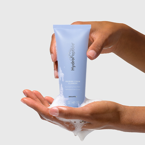 Een paar handen van een model die een lichte paarse tube HydroPeptide Foaming Cream Cleanser vasthouden waar schuim uitkomt