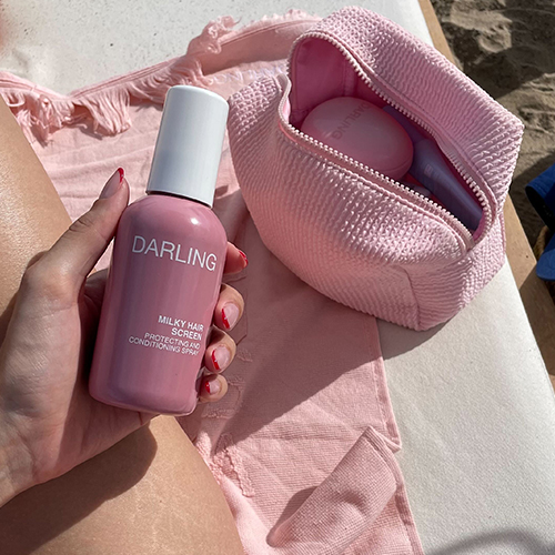 Een hand houdt de roze Darling Milky Hair Screen spray vast op het strand, met een roze toilettas en handdoek op de achtergrond.