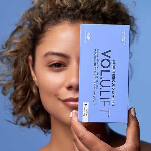 Een lachend model met krullend haar houdt het blauwe doosje van de IMAGE Skincare VOL.U.LIFT creme voor een deel van haar gezicht.
