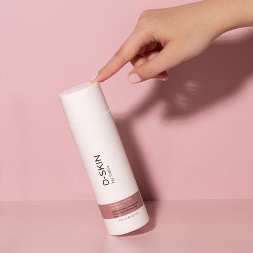 Een hand die de dop aanraakt van een D-SKIN Skin Age Control Cleanser fles tegen een zachte roze achtergrond.