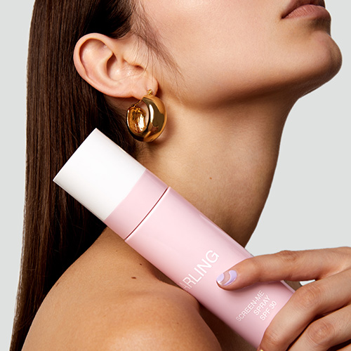 Een model houdt de roze DARLING SCREEN-ME SPF 30 zonnebrandspray tegen haar schouder; ze draagt een gouden oorbel.