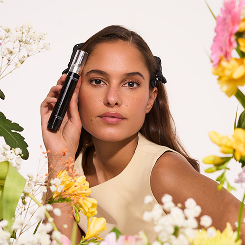 Portret van een vrouw die de Marc Inbane Perle de Soleil tanning drops vasthoudt, omringd door witte en gele bloemen.