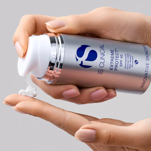 Hand die een witte zonnebrandcrème pompt uit de zilveren flacon van iS Clinical Extreme Protect SPF 40 op de vingers van een andere hand tegen een grijze achtergrond