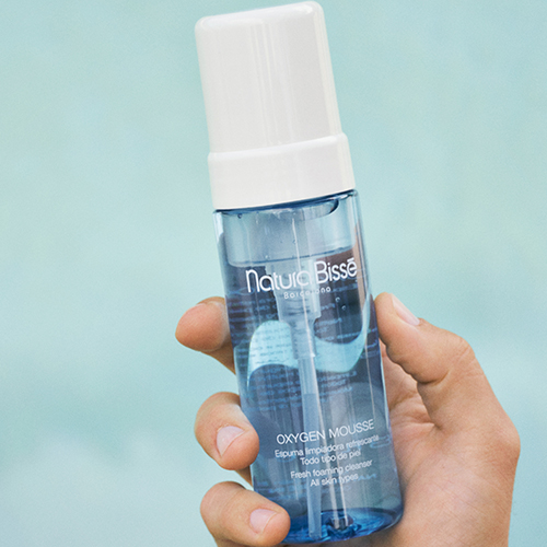 Een hand houdt de blauwe flacon van Natura Bissé Oxygen Mousse vast tegen een heldere, blauwe achtergrond.