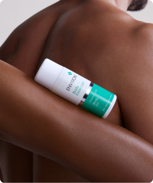 Een sfeerbeeld met afgeronde hoeken waarin een flacon van de Environ Body EssentiA Body Oil rust op de gladde, getinte huid van iemands rug en arm