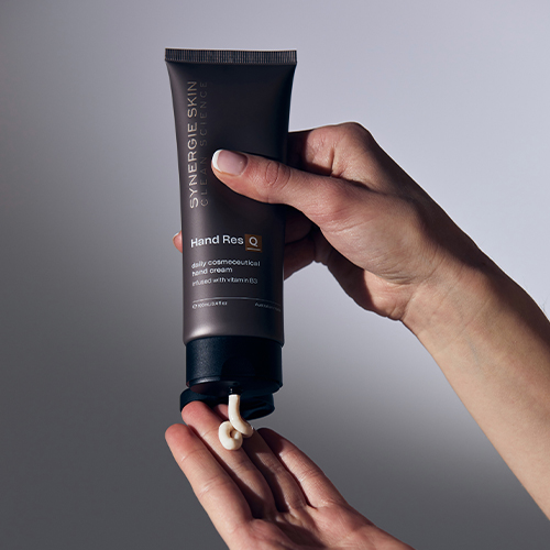 Een model houdt de tube Synergie Skin Hand ResQ handcrème vast en knijpt een kleine hoeveelheid van de witte crème op de andere handpalm om de textuur te tonen.