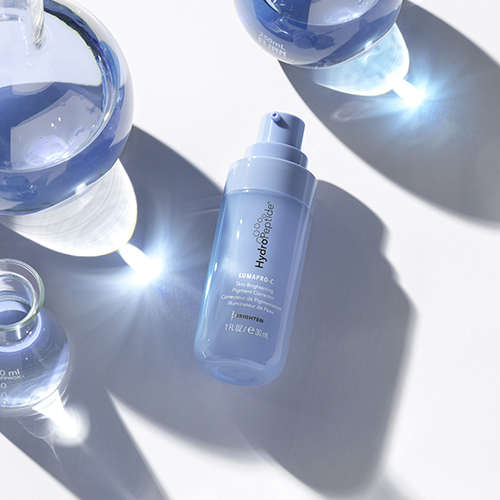 Een blauwe flacon HydroPeptide LumaPro C huidverzorging omringd door glazen laboratoriuminstrumenten en helder licht