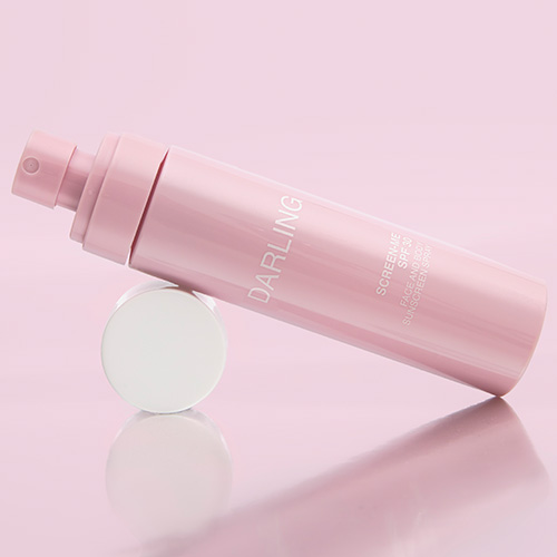 Close-up van een liggende roze DARLING SCREEN-ME SPF 30 flacon met de dop eraf, waarbij de spraykop zichtbaar is.