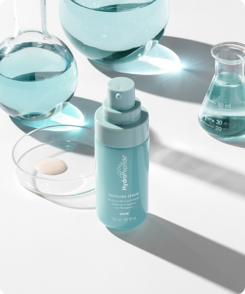 Een lichtblauwe flacon HydroPeptide Soothing Serum op een witte achtergrond tussen glazen laboratoriumkolven met heldere vloeistof en een petrischaaltje met een druppel serum.