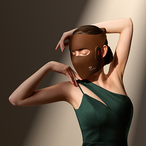 Een vrouw in een groene jurk die het bruine JOVS lichttherapie gezichtsmasker draagt terwijl ze met gesloten ogen poseert in een sfeervolle lichtstraal