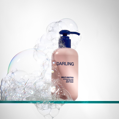 Een dynamische studio-opname op een glazen plank, waar de lichtroze fles Darling Multi-Biotics Body Wash met zijn blauwe pomp omgeven is door een weelderige wolk van doorzichtige zeepbellen. De achtergrond is lichtgrijs met een subtiel verloop.