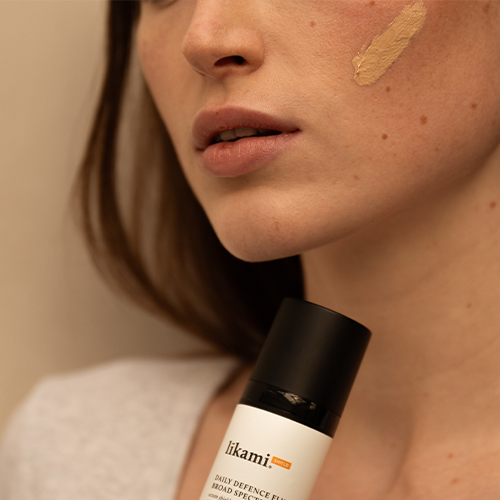 Close-up van de wang van een model waarop een veeg van de getinte Likami Daily Defence Fluid zichtbaar is, met de flacon op de voorgrond.