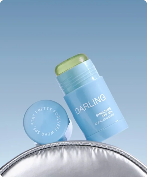 Een geopende DARLING Shield-Me SPF 50+ Clear Sun Stick met een transparante groene textuur, rustend op een zilverkleurige metallic tas tegen een blauwe achtergrond.