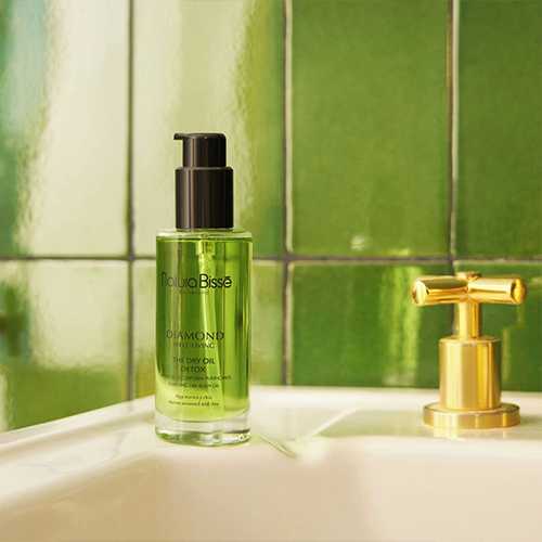 De groene Natura Bissé Detox Dry Body Oil op de rand van een witte wastafel tegen een achtergrond van glanzende groene tegels.
