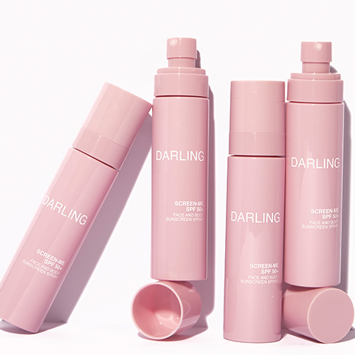 Vier roze flacons van DARLING SCREEN-ME SPF 50+ zonnebrandspray, waarvan twee zonder dop om de vernevelaar te tonen.