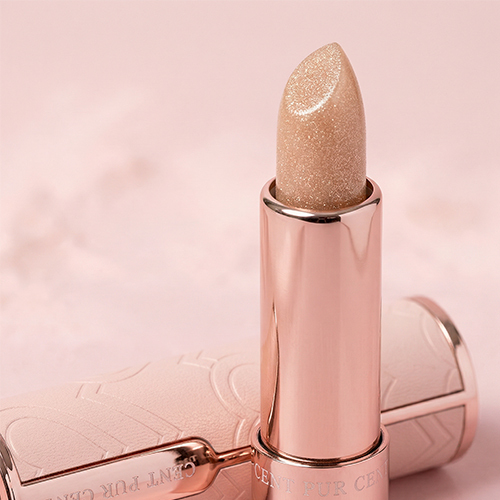 Een close-upfoto van de textuur van een geopende Cent Pur Cent-lippenstift, met een glinsterende, goudkleurige lippenbalsem. De lippenstift staat rechtop op een roze achtergrond en de dop ligt op zijn zijkant.
