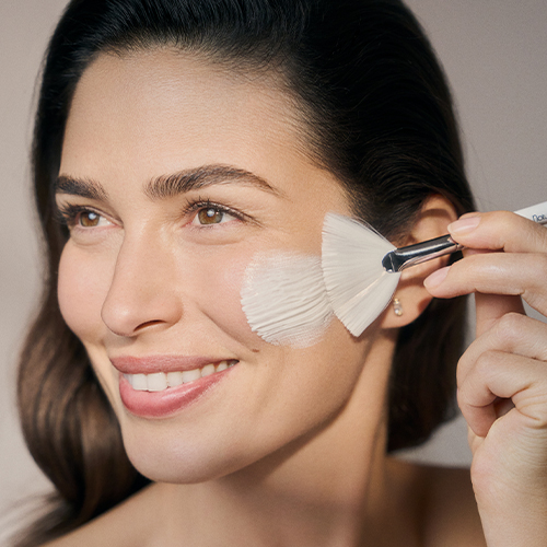 Een close-up van een vrouw die een masker op haar wang aanbrengt met een waaierkwast en glimlacht.