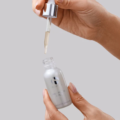 Handen die een mat wit glazen pipetflesje van iS Clinical Brightening Serum vasthouden terwijl er een druppel uit de glazen pipet komt tegen een grijze achtergrond