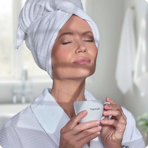 Een vrouw in een witte badjas geniet van een moment van rust met een wit hannah-kopje in een lichte badkameromgeving.