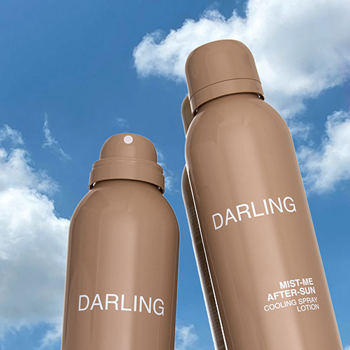 Twee beige spuitbussen van de Darling Mist Me After-Sun cooling spray gefotografeerd tegen een helderblauwe lucht met witte wolken.