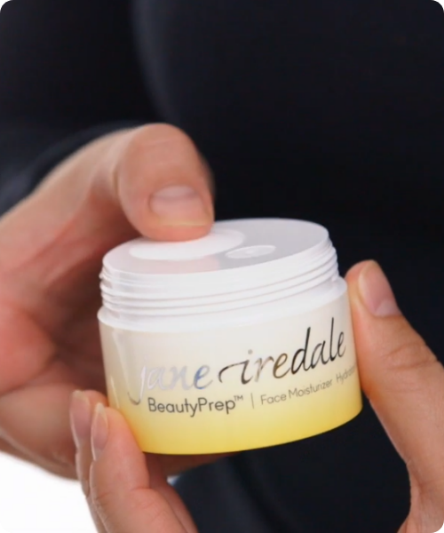 Close-up van handen die de doseerplaat van een Jane Iredale BeautyPrep Face Moisturizer potje indrukken om de crème te doseren.