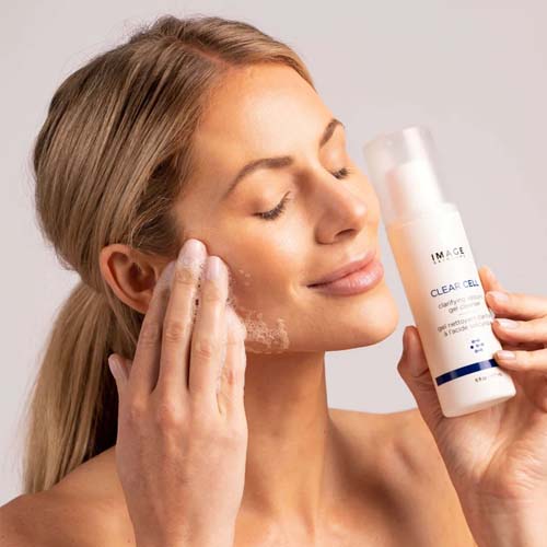 Een glimlachende vrouw reinigt haar gezicht met het schuim van de Clear Cell Gel Cleanser terwijl ze de flacon vasthoudt.