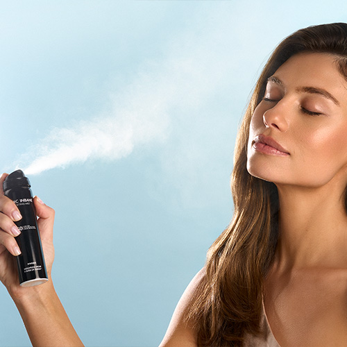 Een model sluit haar ogen terwijl ze de Marc Inbane SPF 50 Setting Mist op haar gezicht vernevelt tegen een blauwe achtergrond
