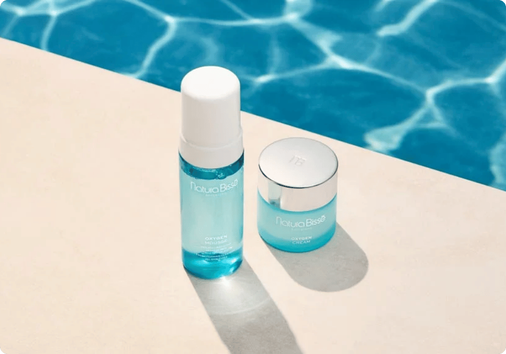 Een flesje Natura Bissé Oxygen Mousse en een potje Oxygen Cream staan naast elkaar op een stenen rand in de zon. Op de achtergrond is het helderblauwe, rimpelende water van een zwembad zichtbaar. De producten hebben een frisse, lichtblauwe uitstraling.