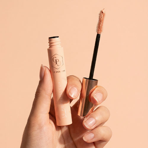 Een hand houdt de geopende Le Merveilleux Lash Primer vast, waarbij het borsteltje met de perzikkleurige textuur zichtbaar is.