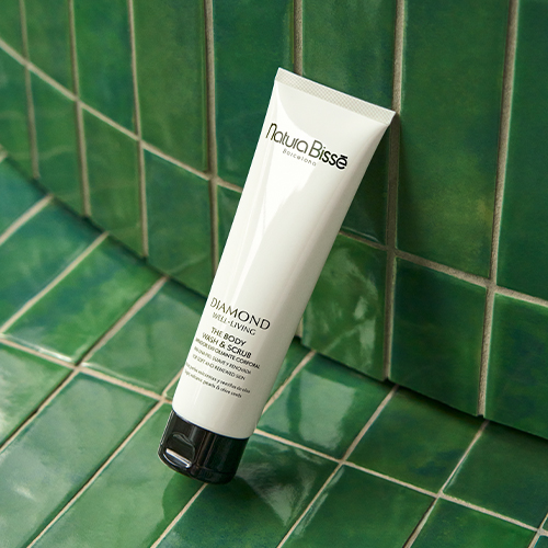 Een vooraanzicht van de Natura Bissé The Body Wash & Scrub tube, die diagonaal rust tegen een hoek van glanzende, smalle, donkergroene metselwerk tegels. De witte tube met zwarte klikdop contrasteert levendig met de tegeltextuur.