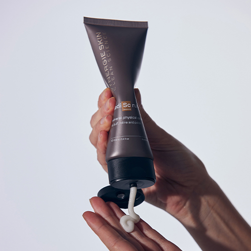 Een detailopname van handen die in de tube Synergie Skin MediScrub knijpen waarbij een egale witte substantie op de vingers wordt aangebracht tegen een witte achtergrond.