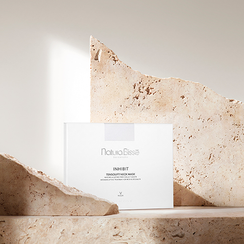 De witte verpakking van het Natura Bissé Inhibit Tensolift Neck Mask, gepresenteerd in een luxe setting tussen natuurlijke beige stenen en zacht invallend zonlicht.
