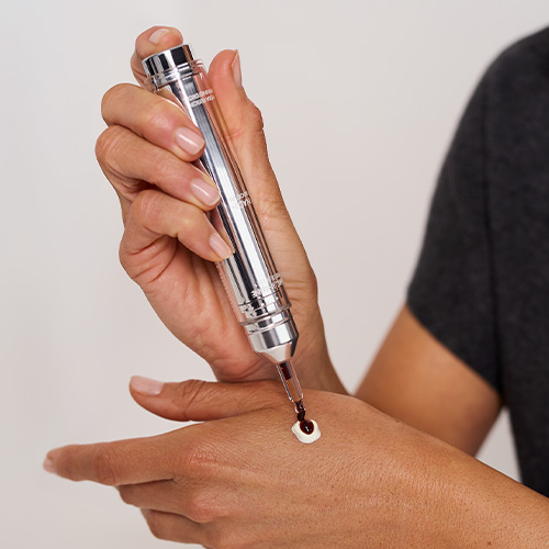 Close-up van een hand waarop een druppel donkerbruin tanning-serum wordt toegevoegd aan een toefje witte gezichtscrème met de precisie-applicator.