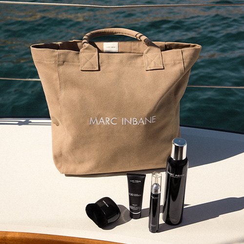De beige Marc Inbane canvas tas staat op het dek van een boot bij het water met daarvoor verschillende luxe zelfbruiner en zonnebrandproducten van het merk.