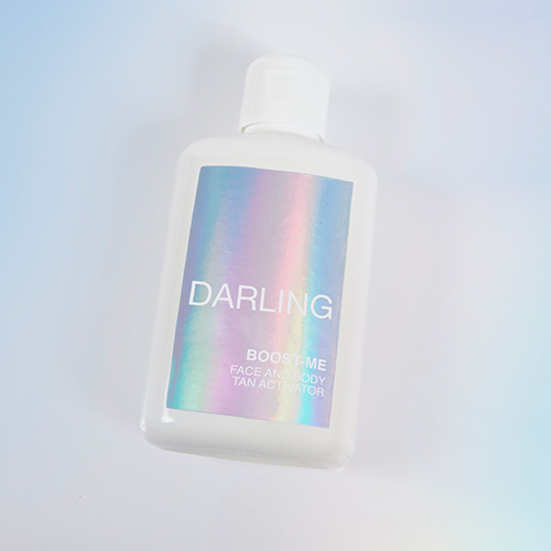 Een schuine weergave van de DARLING BOOST-ME Tan Activator flacon op een lichte ondergrond met zachte pastelkleurige reflecties.